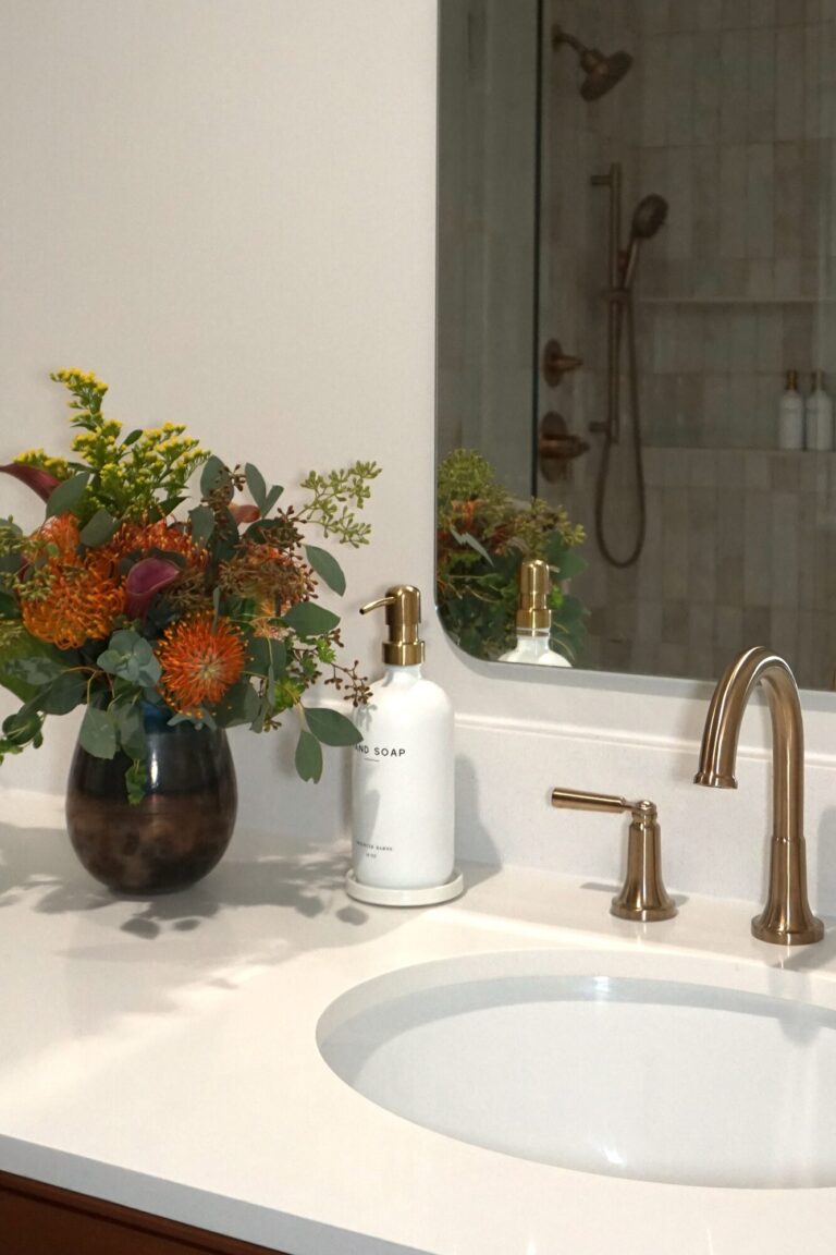 Close up of a custom bathroom vanity with a white countertop, brushed brass faucet, arched mirror, and floral arrangement for a warm, modern look.