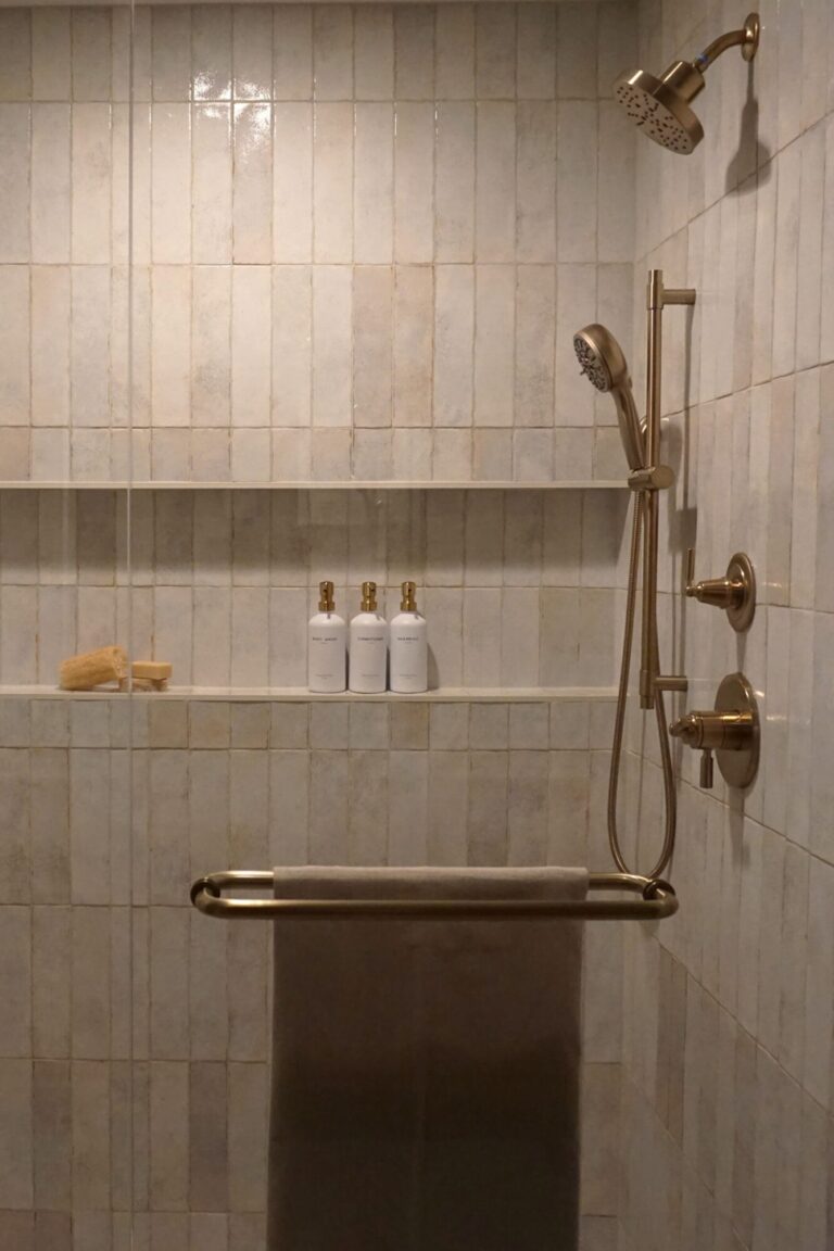 Walk in shower with vertical neutral tile, built in shelf, brushed brass fixtures, and minimalist bath accessories.