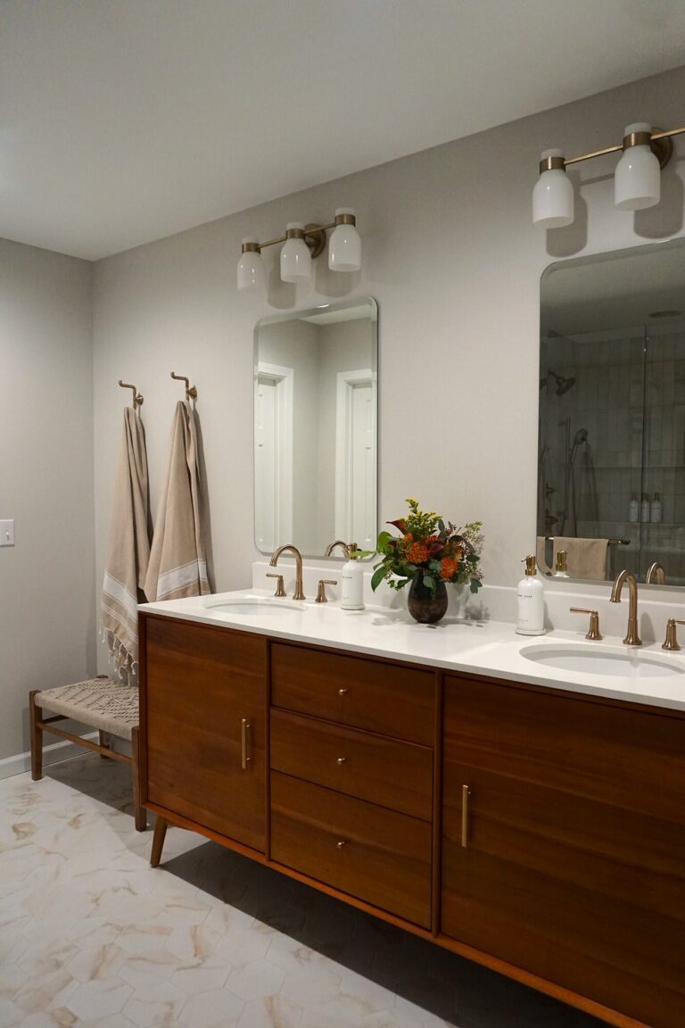 Double sink bathroom vanity with warm wood cabinetry, white countertop, brass fixtures, and soft neutral finishes.