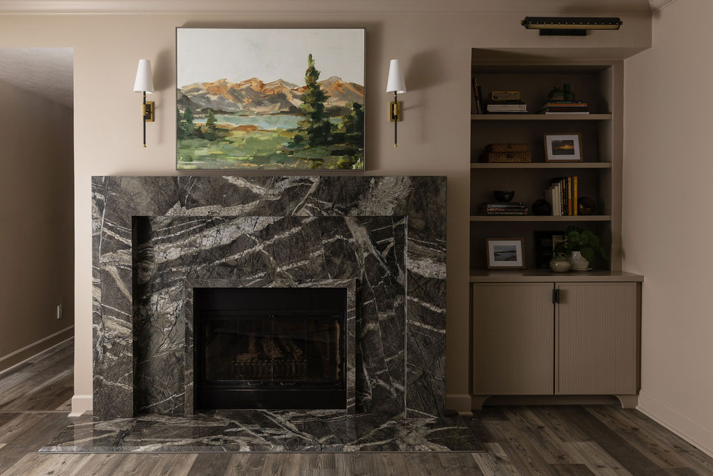 Dark marble fireplace with a landscape painting above the mantel, flanked by brass sconces and open wood shelving.