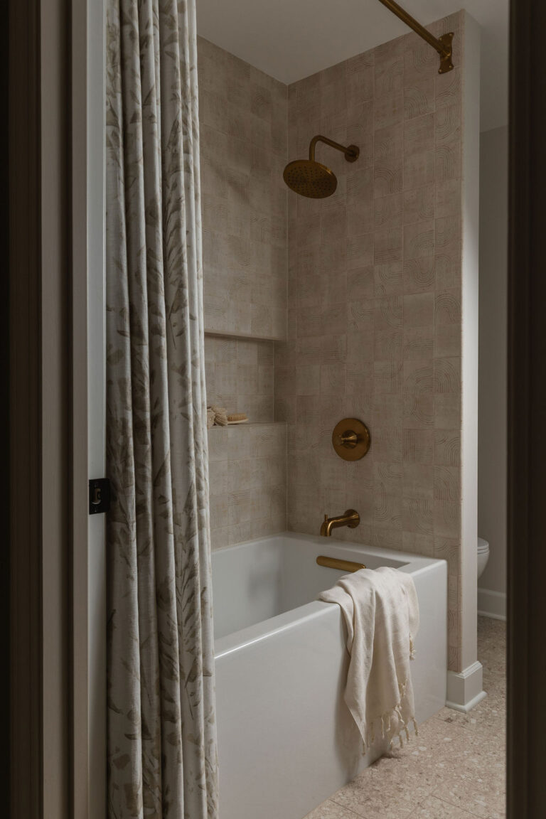 Soaking tub with striped curtain and brass fixtures set against textured tan tile.