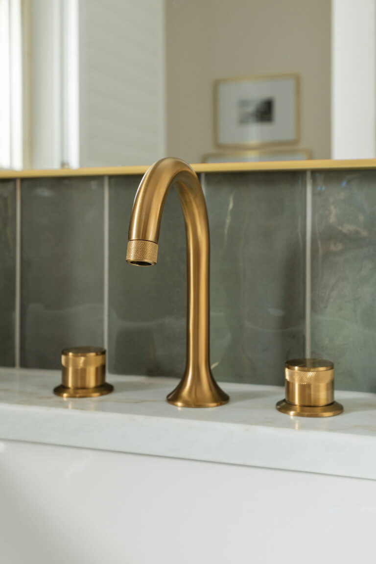 Close-up of a brass widespread faucet against a white marble countertop and green tile backsplash.