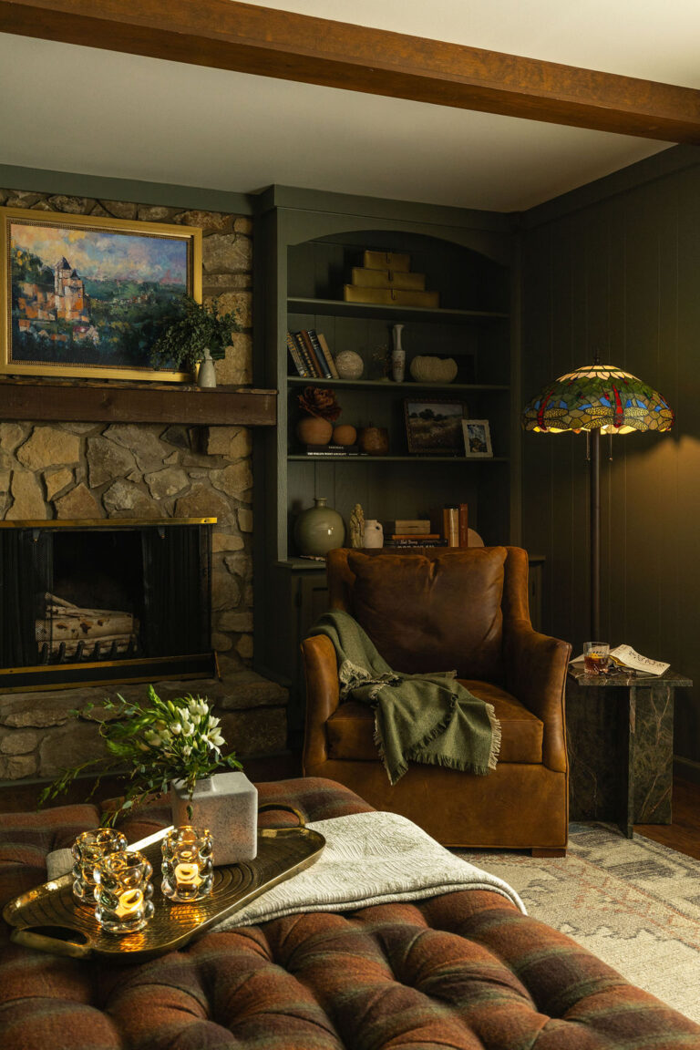Cognac leather wingback chair with a fringed olive throw beside a stone fireplace and built-in bookshelf, lit by a stained glass floor lamp.
