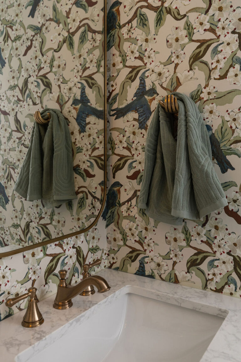 Sage green towels hanging on brass hooks against botanical bird print wallpaper.