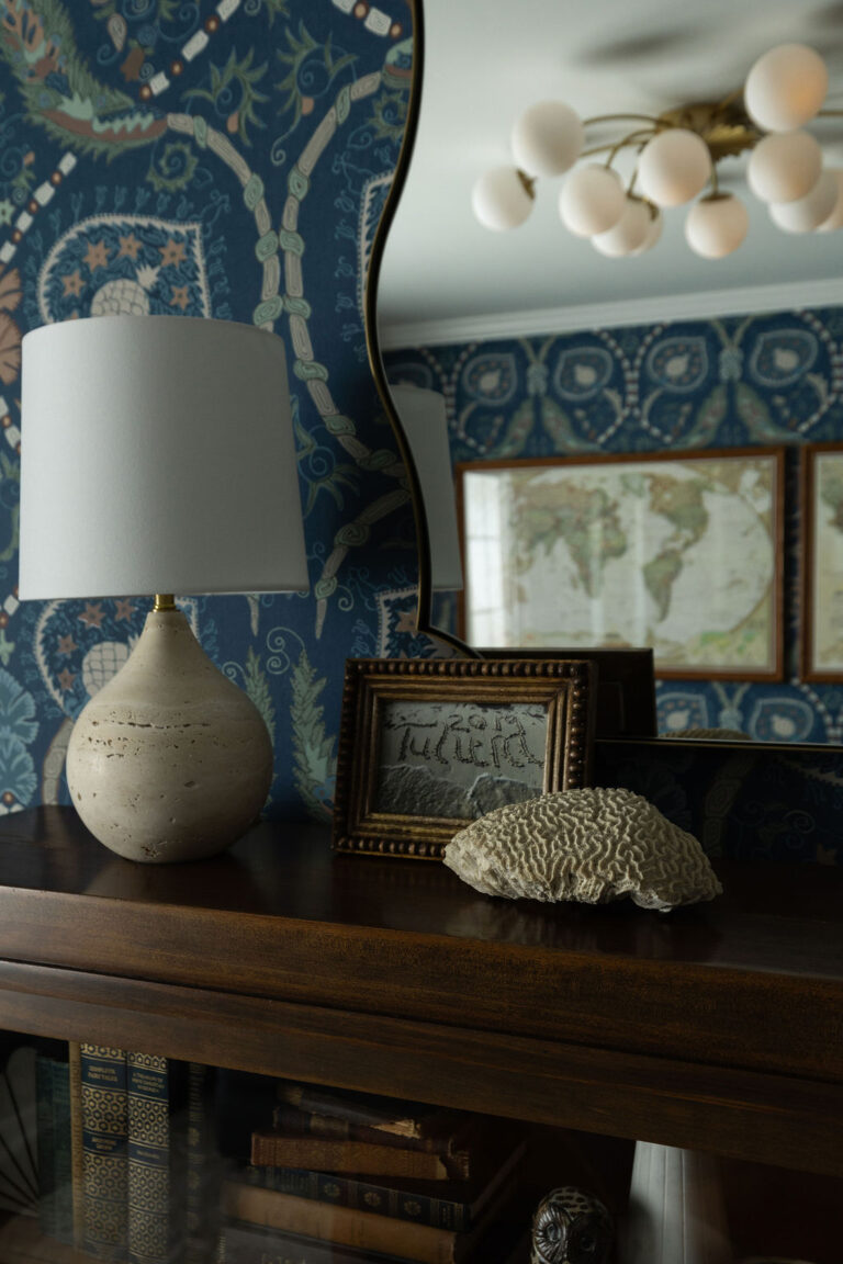 Dark wood dresser with a ceramic lamp, framed map, and a piece of natural coral against blue patterned wallpaper.