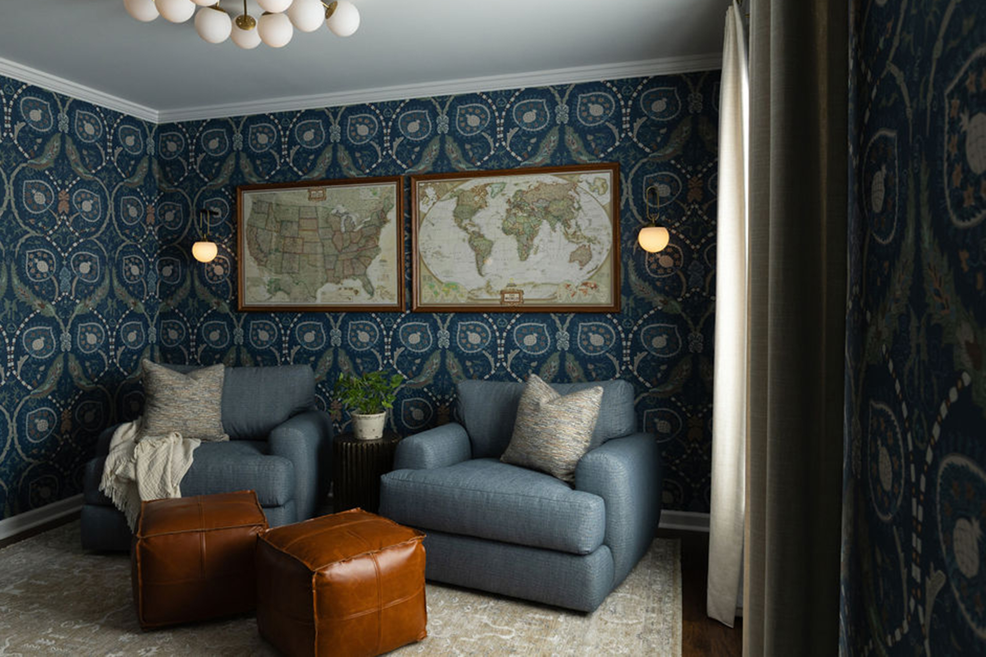 Blue patterned wallpaper sitting room with two matching armchairs, leather cube ottomans, and framed vintage maps flanking pendant sconces.