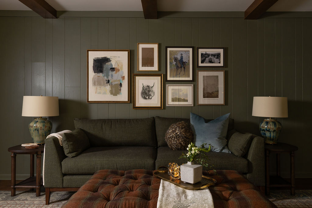 Deep olive green living room with a gallery wall of framed art above a dark green sofa, flanked by ceramic table lamps and a plaid tufted ottoman.