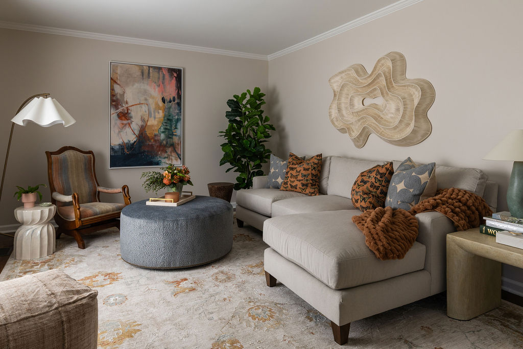 Neutral living room with a sectional sofa, round blue ottoman, and layered abstract art above a leafy potted plant.